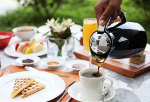 mesa de café da manhã com xícara de café com café sendo servido, torradas, tortas, suco em um espaço de jardim do restaurante Tano