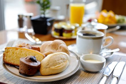mesa de café da manhã com prato cheio de pães diferentes e uma xícara de café ao lado