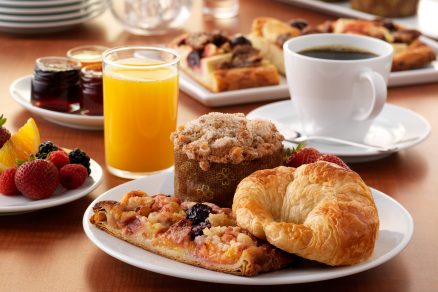 mesa com refeições matinais como pães, torradas, um copo com suco, uma xícara com café representando o café da manhã pelo mundo