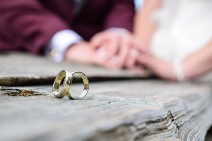 Duas alianças em foco com mãos de casal ao fundo.