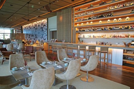 Salão sofisticado de restaurante no Grand Hyatt Rio de Janeiro, com bar iluminado ao fundo repleto de garrafas e chapéus decorativos, mesas com poltronas estofadas claras, piso de madeira e ambiente contemporâneo elegante.