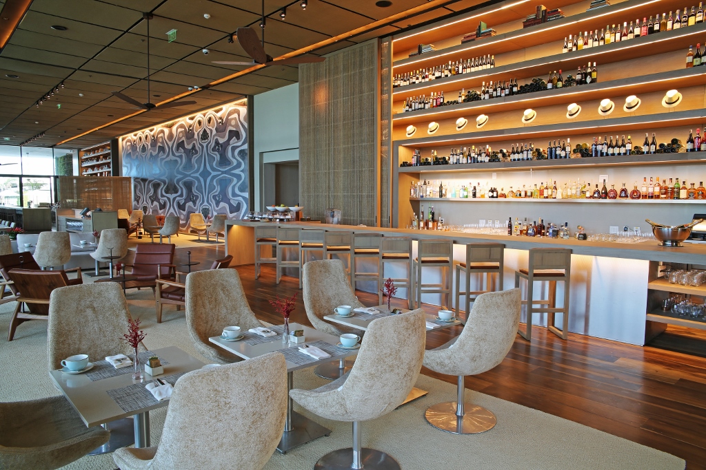 Salão sofisticado de restaurante no Grand Hyatt Rio de Janeiro, com bar iluminado ao fundo repleto de garrafas e chapéus decorativos, mesas com poltronas estofadas claras, piso de madeira e ambiente contemporâneo elegante.