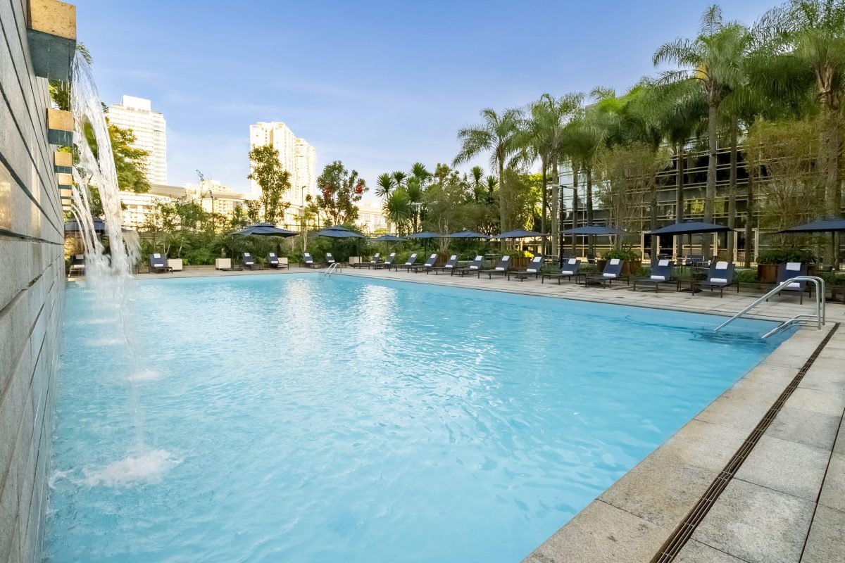 Piscina externa de hotel de luxo em São Paulo, com água azul cristalina, queda d’água lateral, espreguiçadeiras alinhadas com guarda-sóis, palmeiras ao redor e prédios ao fundo sob céu ensolarado.
