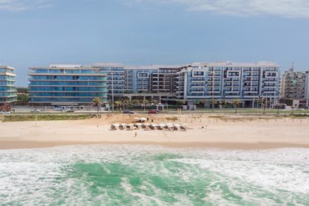 Fachada do Grand Hyatt Rio de Janeiro vista da praia da Barra da Tijuca, com mar em tons de verde, faixa de areia com ombrelones organizados e o hotel de luxo ao fundo em um dia ensolarado.