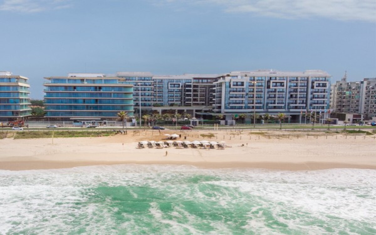 Fachada do Grand Hyatt Rio de Janeiro vista da praia da Barra da Tijuca, com mar em tons de verde, faixa de areia com ombrelones organizados e o hotel de luxo ao fundo em um dia ensolarado.